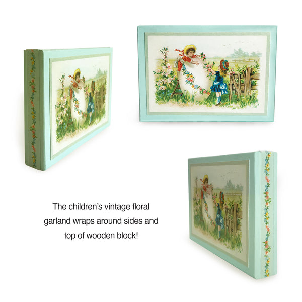 Elegant vintage-inspired Easter wood block made with end-of-19th-century postcard. Decorate your home or gift to a friend this elegant reminder of what it was like to send Easter postcards to relatives and friends! Reproduced vintage “Easter Greetings” postcard. Easter Vintage Decorations. Elegant illustration features young girl and boy decorating an Easter egg in the meadow. Easter egg garland on postcard used to create elegant border on sides of wood block. tribegift.com