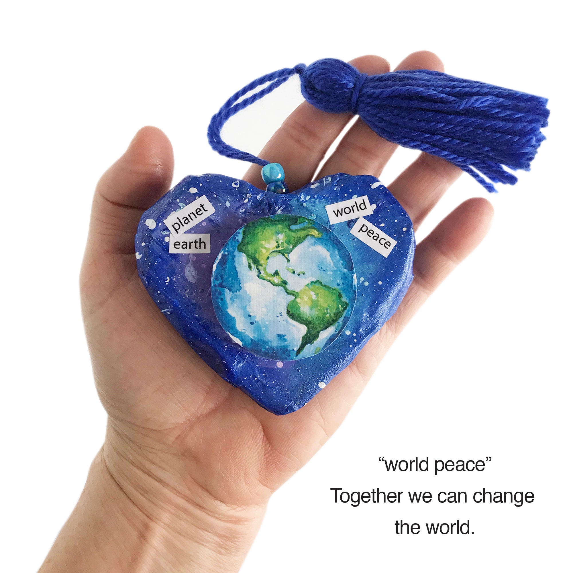 Heart paper mache sculpture celebrates planet earth and world peace. Perfect peace gift uniquely created to fit in palm of hand. One-of-a-kind hand sculpted and hand-painted shape. Earth divided into two halves with hemisphere on each side of heart. Color selection promotes sustainable global awareness that encourages us to respect, protect and preserve planet earth and the cosmos. Earth illustration suspends up from outer-space-colored background. Deep blue yarn tassel attached. Tribegift.com